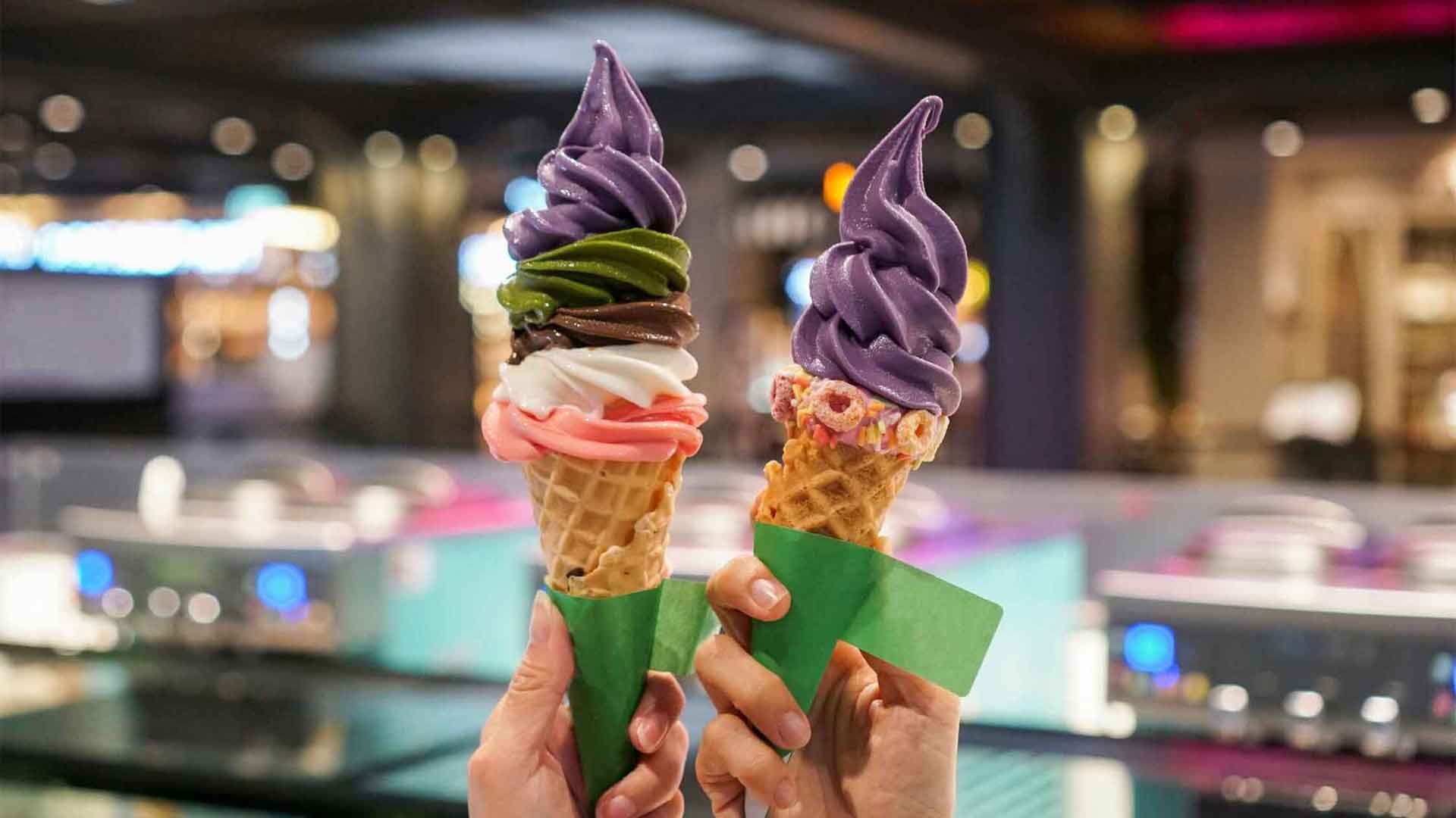 Two ice cream cones with colorful swirls held against a blurred indoor background.