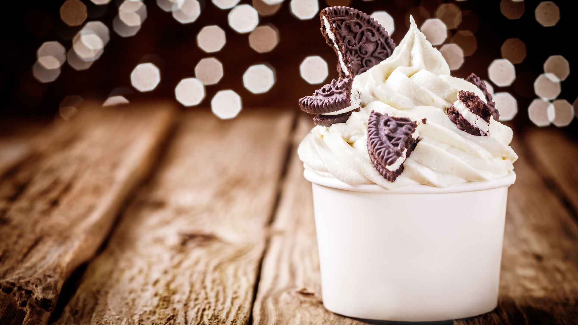 Cupcake with white frosting and chocolate cookies on a wooden surface with blurred lights in the background
