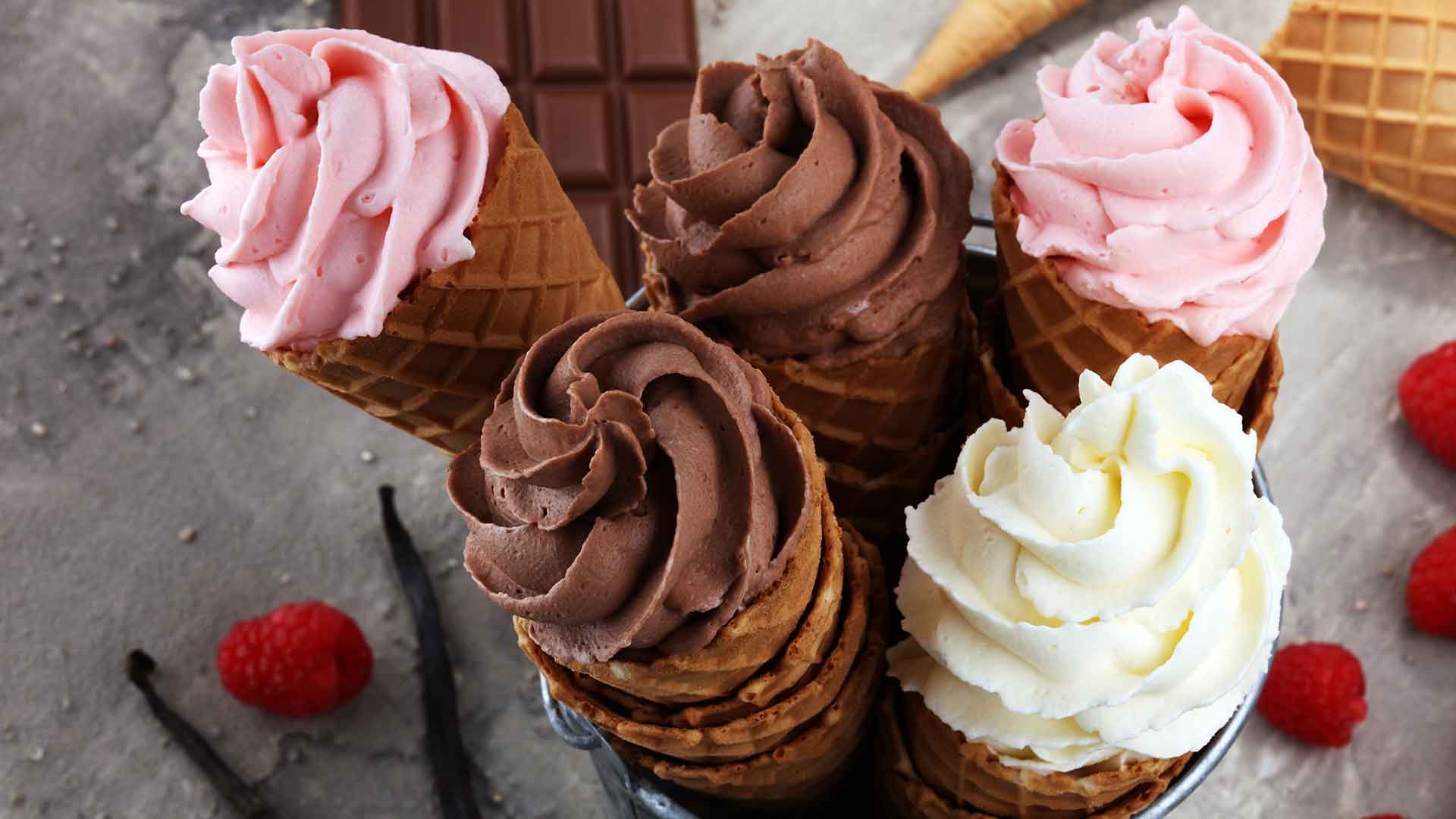 Ice cream cones with pink, chocolate, and white frosting on a gray surface with raspberries and a vanilla bean.