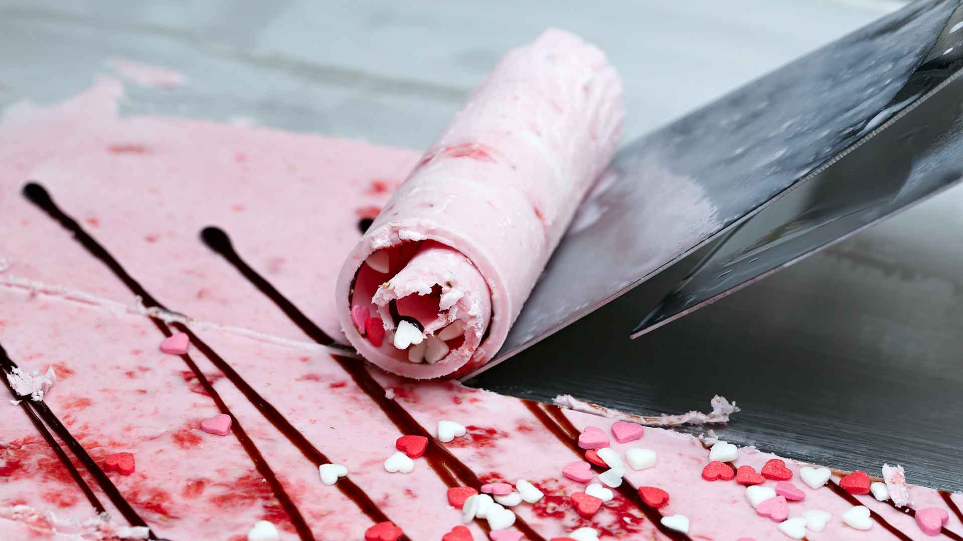 Close-up of a rolled dessert with pink and white filling, drizzled with red sauce and sprinkled with red and white candies on a reflective surface.