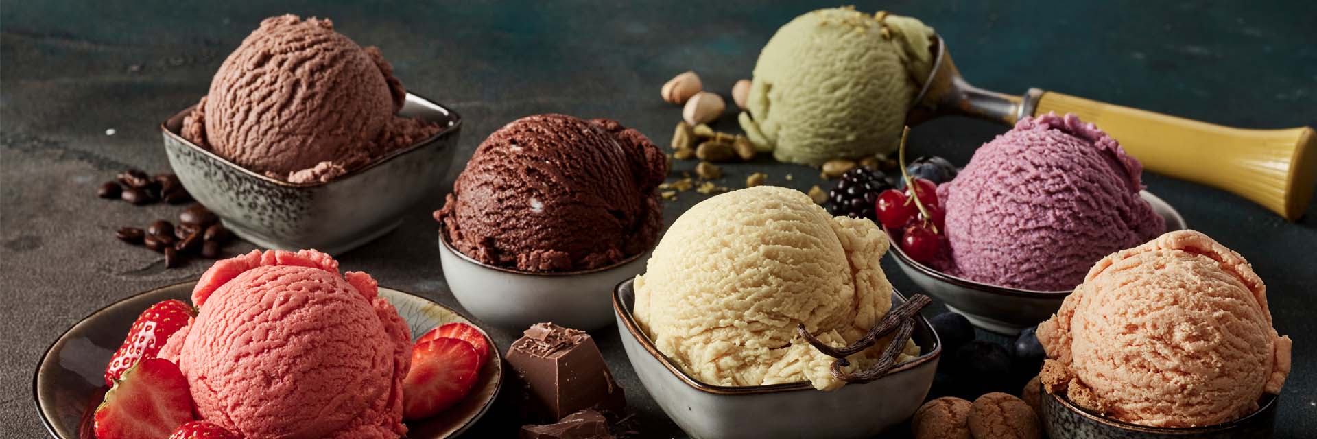 Assorted ice cream scoops in bowls on a dark surface with berries and chocolate pieces.
