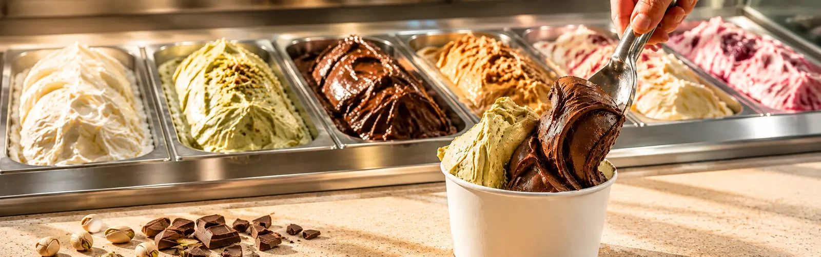Ice cream display with various flavors and a cup of ice cream on a wooden surface.